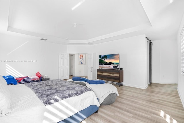 bedroom with a barn door, visible vents, baseboards, light wood finished floors, and a raised ceiling