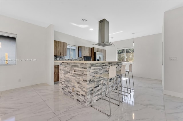 kitchen with a breakfast bar, island range hood, stainless steel fridge with ice dispenser, hanging light fixtures, and decorative backsplash