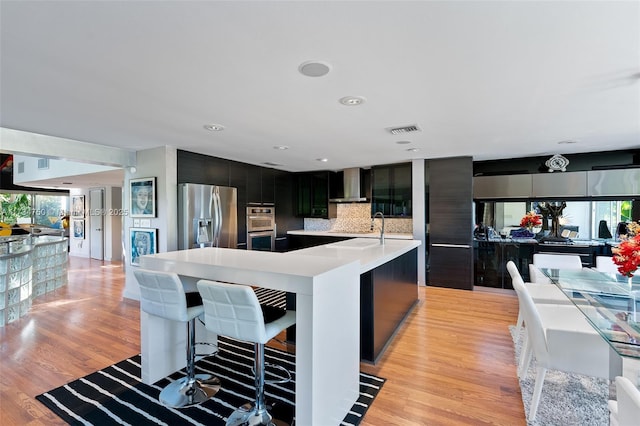 kitchen featuring an island with sink, a breakfast bar area, stainless steel appliances, wall chimney range hood, and light wood-type flooring