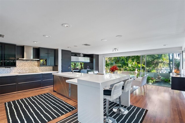 kitchen with wall chimney range hood, a kitchen island with sink, tasteful backsplash, light hardwood / wood-style floors, and a kitchen bar