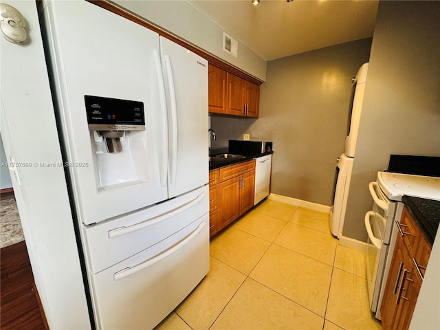 kitchen with white appliances, light tile patterned floors, dark countertops, brown cabinets, and a sink