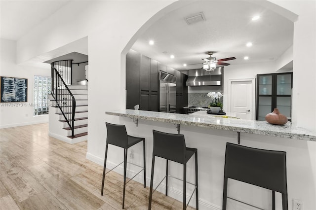 kitchen featuring wall chimney range hood, ceiling fan, light stone counters, kitchen peninsula, and stainless steel built in fridge