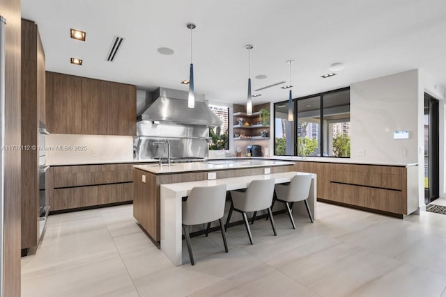 kitchen with hanging light fixtures, a kitchen island with sink, ventilation hood, light countertops, and open shelves