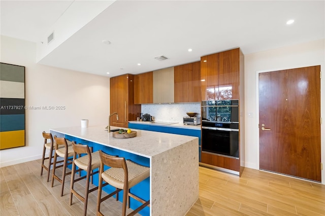 kitchen featuring light hardwood / wood-style flooring, a kitchen breakfast bar, a kitchen island with sink, decorative backsplash, and black appliances