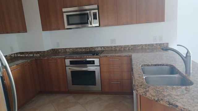 kitchen featuring light tile patterned floors, appliances with stainless steel finishes, brown cabinetry, a sink, and light stone countertops