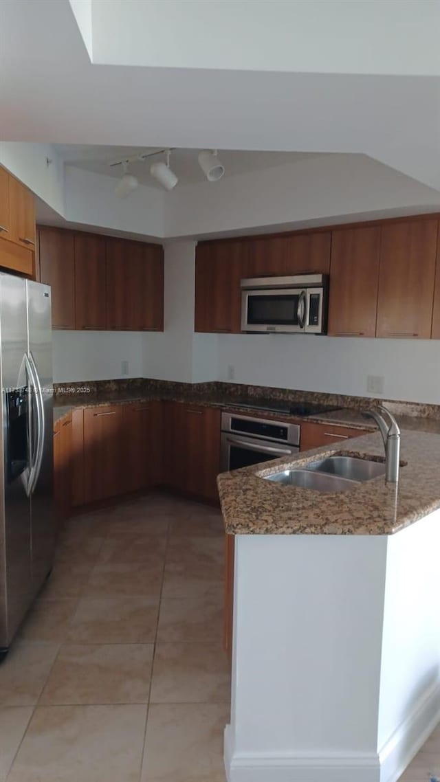 kitchen with stainless steel appliances, a peninsula, a sink, brown cabinets, and dark stone counters