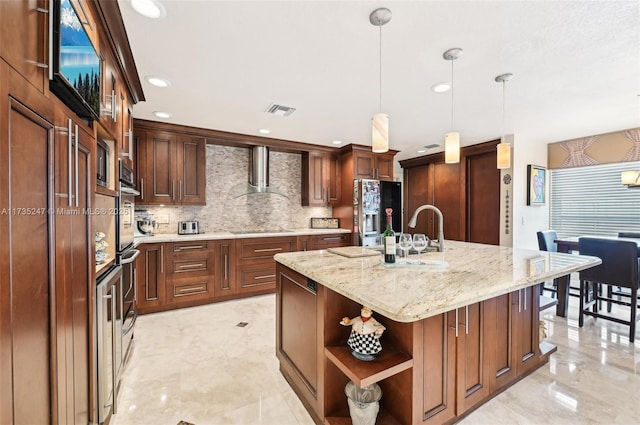 kitchen with wall chimney exhaust hood, a center island with sink, appliances with stainless steel finishes, pendant lighting, and light stone countertops