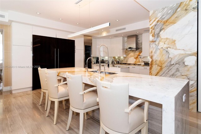 kitchen featuring a breakfast bar, white cabinets, wall chimney exhaust hood, light stone countertops, and a spacious island