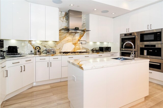 kitchen with wall chimney exhaust hood, white cabinetry, a center island with sink, appliances with stainless steel finishes, and light stone countertops