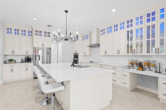 kitchen with wall chimney exhaust hood, sink, light stone counters, stainless steel appliances, and a kitchen island with sink