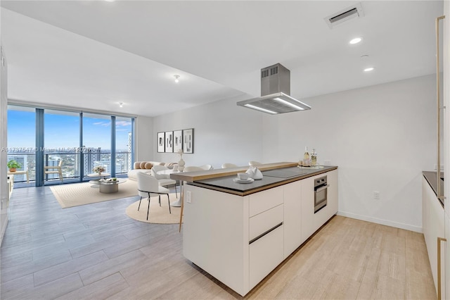 kitchen featuring white cabinets, expansive windows, island exhaust hood, stainless steel oven, and light hardwood / wood-style flooring