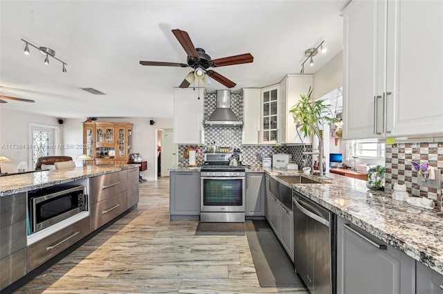 kitchen with ceiling fan, appliances with stainless steel finishes, white cabinetry, light stone counters, and wall chimney exhaust hood