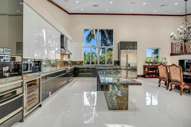 kitchen with beverage cooler, ornamental molding, white cabinetry, appliances with stainless steel finishes, and wall chimney range hood