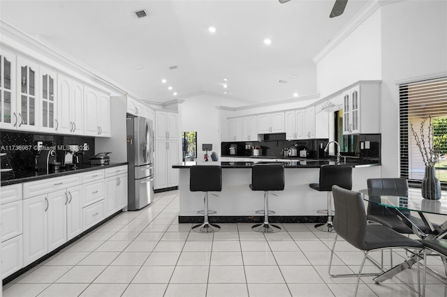 kitchen with stainless steel refrigerator with ice dispenser, dark countertops, glass insert cabinets, ornamental molding, and a kitchen bar