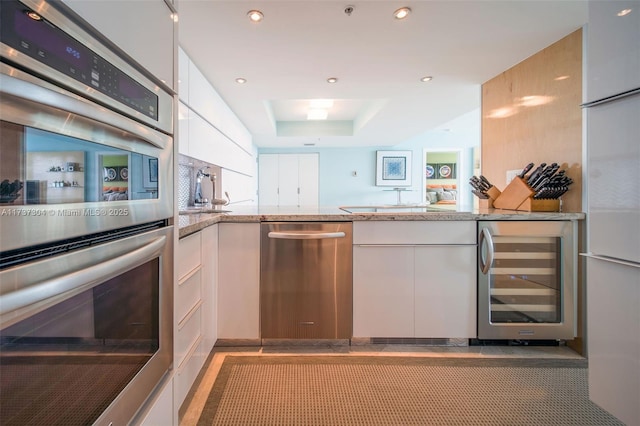 kitchen with appliances with stainless steel finishes, white cabinetry, wine cooler, light stone counters, and a tray ceiling