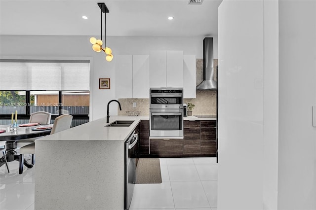 kitchen featuring wall chimney range hood, sink, hanging light fixtures, stainless steel appliances, and white cabinets