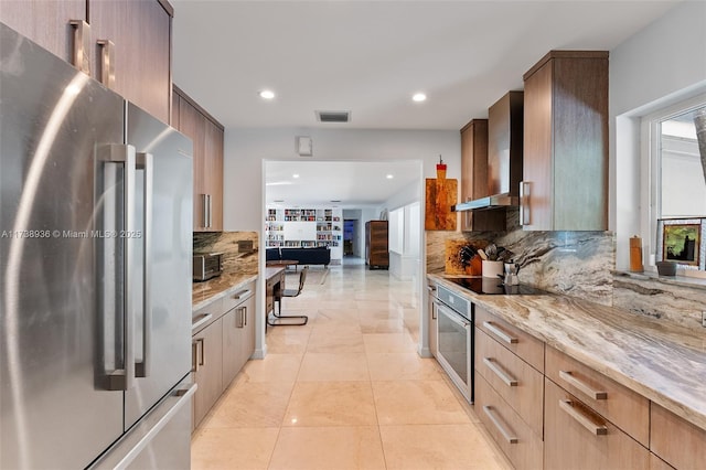 kitchen with appliances with stainless steel finishes, decorative backsplash, light tile patterned floors, light stone countertops, and wall chimney exhaust hood