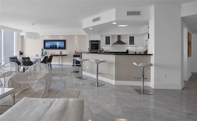 kitchen with double oven, white cabinetry, a kitchen bar, kitchen peninsula, and wall chimney range hood