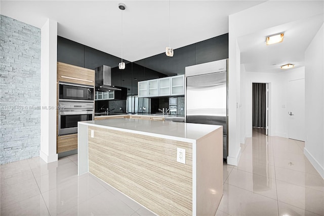 kitchen with wall chimney exhaust hood, built in appliances, decorative light fixtures, light tile patterned floors, and an island with sink