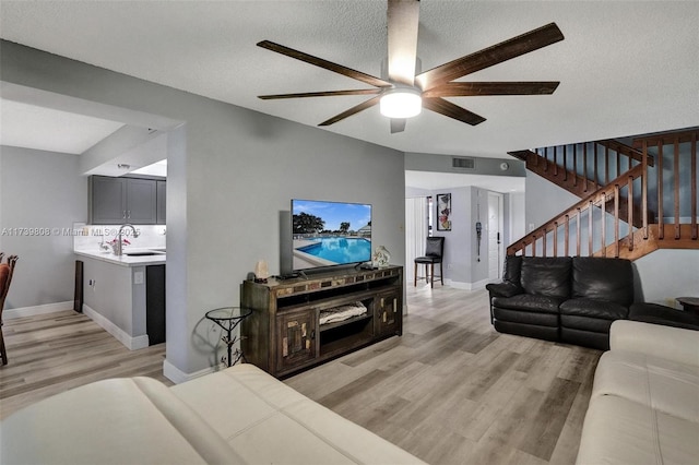 living room featuring stairs, light wood-style floors, visible vents, a ceiling fan, and a textured ceiling