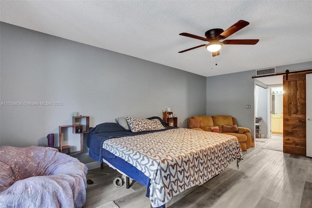 bedroom featuring a textured ceiling, visible vents, a barn door, wood finished floors, and a ceiling fan
