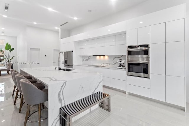 kitchen with sink, a center island with sink, stainless steel double oven, decorative backsplash, and white cabinets