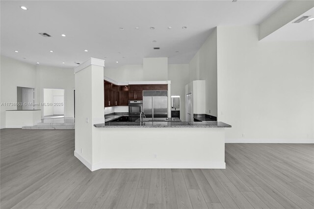 kitchen with dark brown cabinetry, kitchen peninsula, dark stone countertops, and stainless steel built in refrigerator