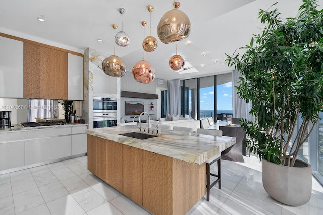 kitchen with white cabinetry, sink, stainless steel double oven, hanging light fixtures, and a center island with sink