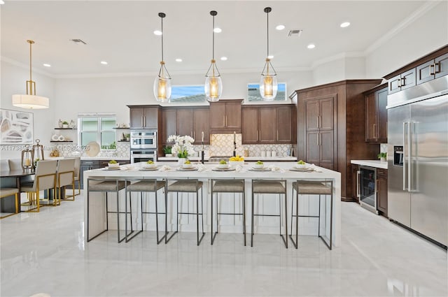 kitchen with dark brown cabinets, hanging light fixtures, a center island with sink, stainless steel appliances, and beverage cooler