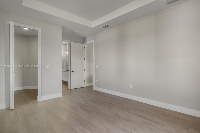 unfurnished bedroom with ensuite bath, a spacious closet, light wood-type flooring, and a tray ceiling