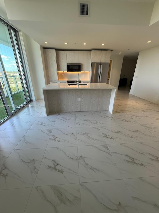 kitchen with sink, a wall of windows, a kitchen island with sink, stainless steel appliances, and light brown cabinets