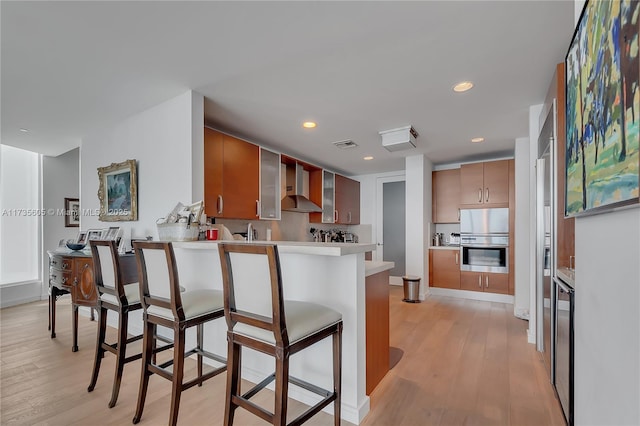 kitchen with a breakfast bar area, backsplash, light hardwood / wood-style floors, kitchen peninsula, and wall chimney exhaust hood