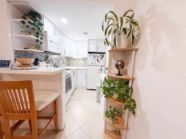 kitchen featuring a breakfast bar area, light tile patterned floors, white cabinets, white appliances, and backsplash