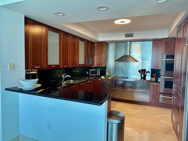 kitchen with double oven, stovetop, decorative backsplash, kitchen peninsula, and wall chimney range hood