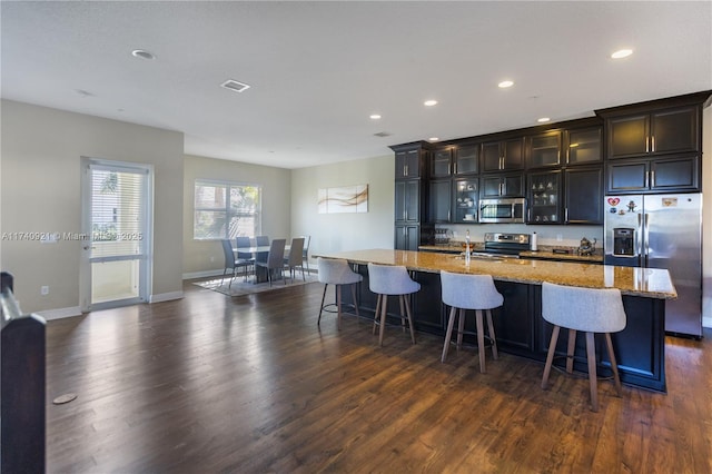 kitchen featuring dark hardwood / wood-style floors, an island with sink, a kitchen bar, stainless steel appliances, and light stone countertops