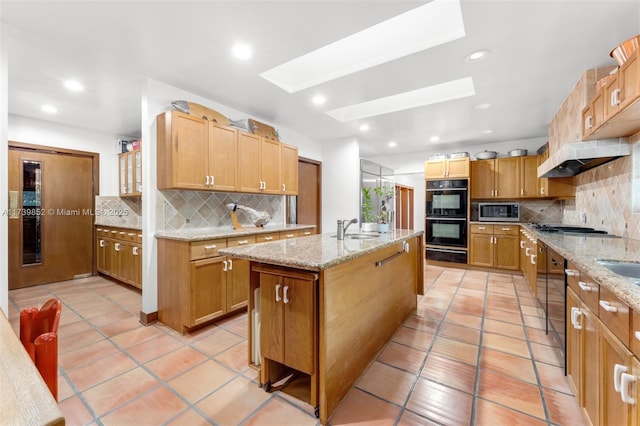 kitchen with a skylight, sink, light stone counters, stainless steel appliances, and a center island with sink