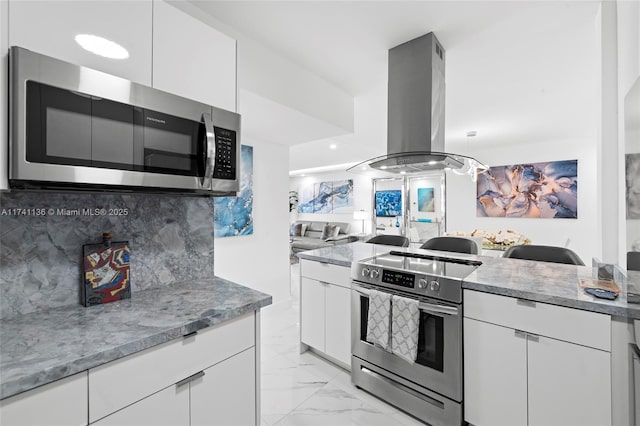 kitchen with island range hood, white cabinetry, marble finish floor, appliances with stainless steel finishes, and decorative backsplash