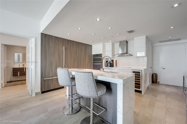 kitchen featuring white cabinetry, beverage cooler, decorative backsplash, a kitchen island with sink, and wall chimney range hood