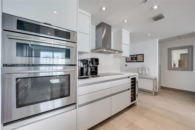 kitchen with wall chimney exhaust hood, tasteful backsplash, double oven, black electric stovetop, and white cabinets