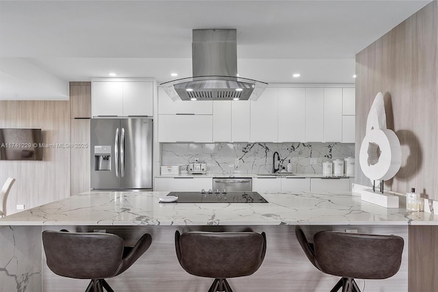 kitchen featuring appliances with stainless steel finishes, white cabinetry, a kitchen breakfast bar, island range hood, and light stone countertops