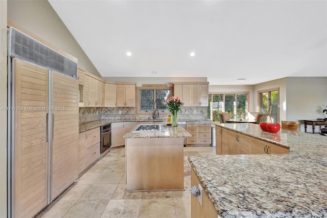 kitchen with appliances with stainless steel finishes, a center island, light brown cabinetry, light stone countertops, and decorative backsplash