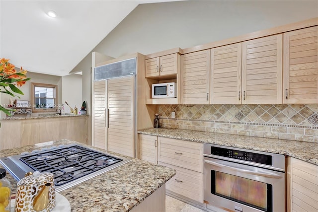 kitchen with stainless steel appliances, vaulted ceiling, light stone counters, light brown cabinets, and decorative backsplash