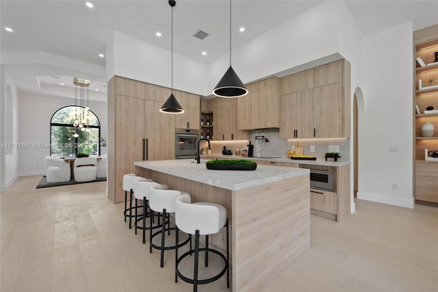 kitchen featuring hanging light fixtures, stainless steel appliances, tasteful backsplash, an island with sink, and light brown cabinets