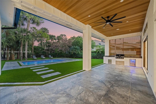 patio terrace at dusk featuring exterior kitchen, a yard, grilling area, ceiling fan, and a fenced in pool