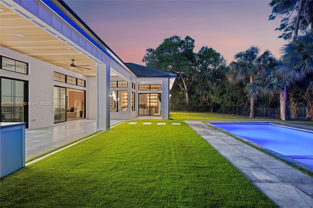 pool at dusk featuring a lawn, ceiling fan, and a patio area