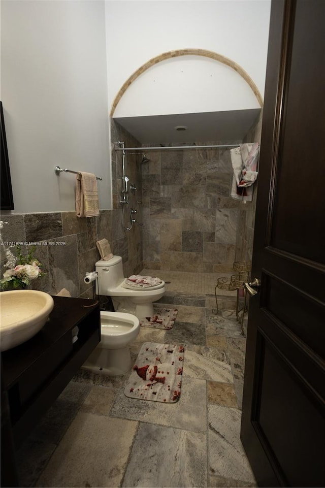 bathroom with a tile shower, tile walls, vanity, toilet, and a bidet