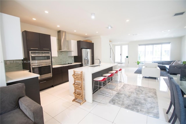 kitchen with sink, tasteful backsplash, a center island with sink, appliances with stainless steel finishes, and wall chimney range hood