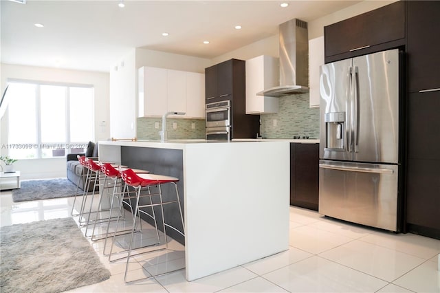 kitchen with wall chimney range hood, white cabinetry, stainless steel appliances, tasteful backsplash, and a kitchen bar