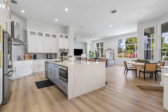 kitchen featuring appliances with stainless steel finishes, white cabinets, a kitchen island with sink, light stone countertops, and wall chimney exhaust hood