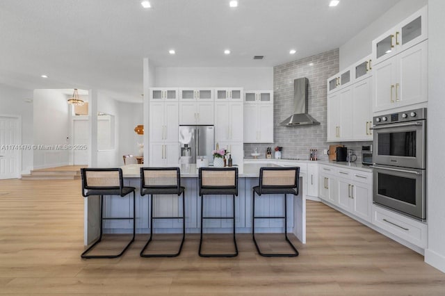 kitchen featuring white cabinetry, stainless steel appliances, an island with sink, and wall chimney range hood